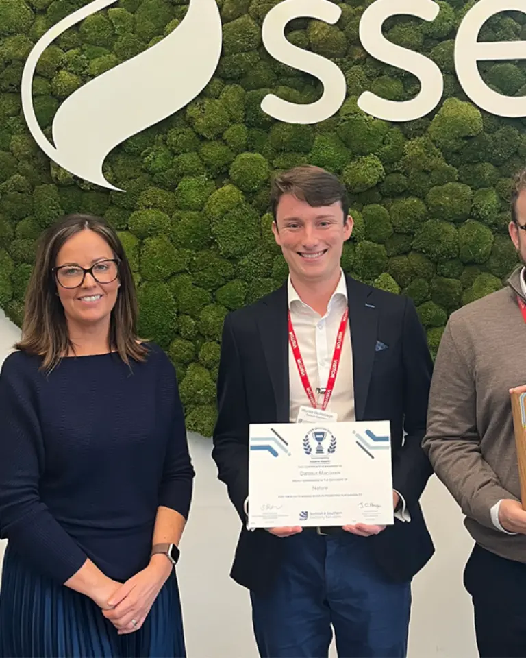 Three individuals standing in front of a moss-covered wall with the logo 'sse' displayed. The middle person is holding a certificate, while another holds a trophy. They are all smiling and wearing lanyards.