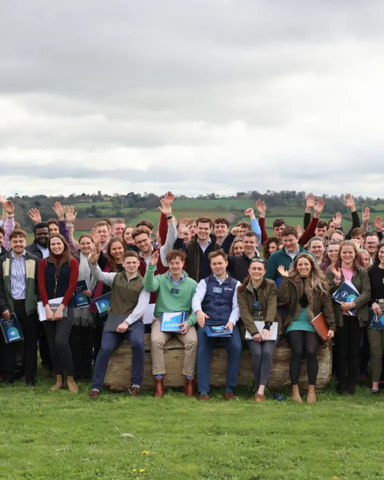 A large group of individuals standing outdoors with their arms raised in celebration. They are dressed in a mix of casual and formal attire, with green fields and overcast skies in the background.