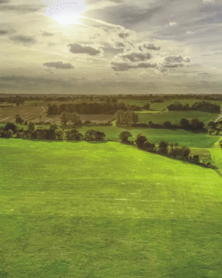 Aerial view of a lush green field bordered by trees, with a highway running alongside it. The sky is partly cloudy, and sunlight is visible, casting a warm glow over the landscape.