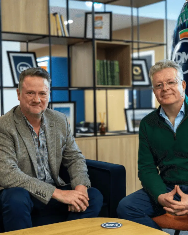 Two men seated on a couch in a modern office setting. One man is wearing a blazer and the other a green sweater. They are positioned around a wooden coffee table, with books and framed pictures in the background.