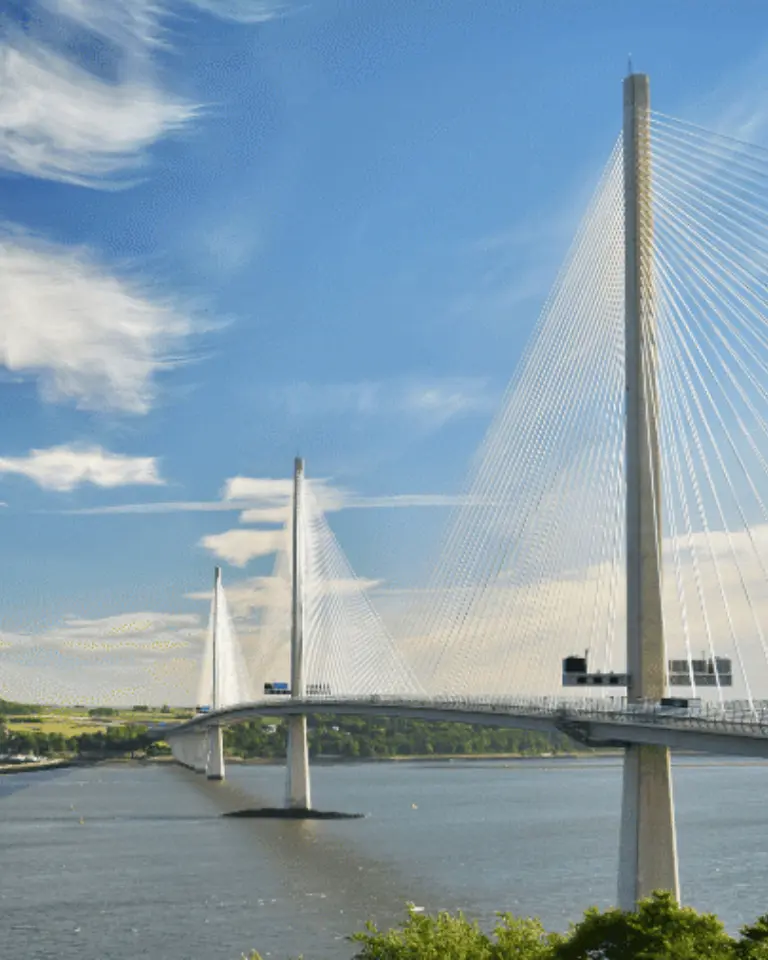 A suspension bridge with tall towers and cable stays spans across a river, beneath a clear blue sky with scattered clouds. Lush greenery is visible in the foreground.