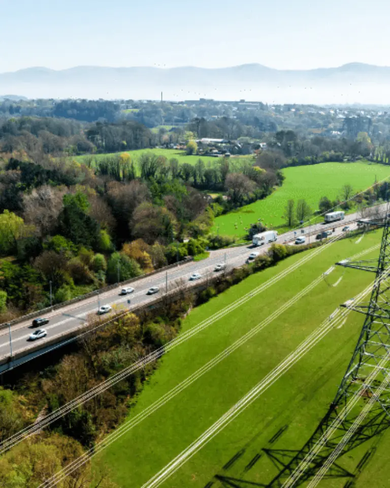 An aerial view of a rural landscape with green fields, a highway with cars, and power lines. Rolling hills are visible in the background under a clear blue sky.