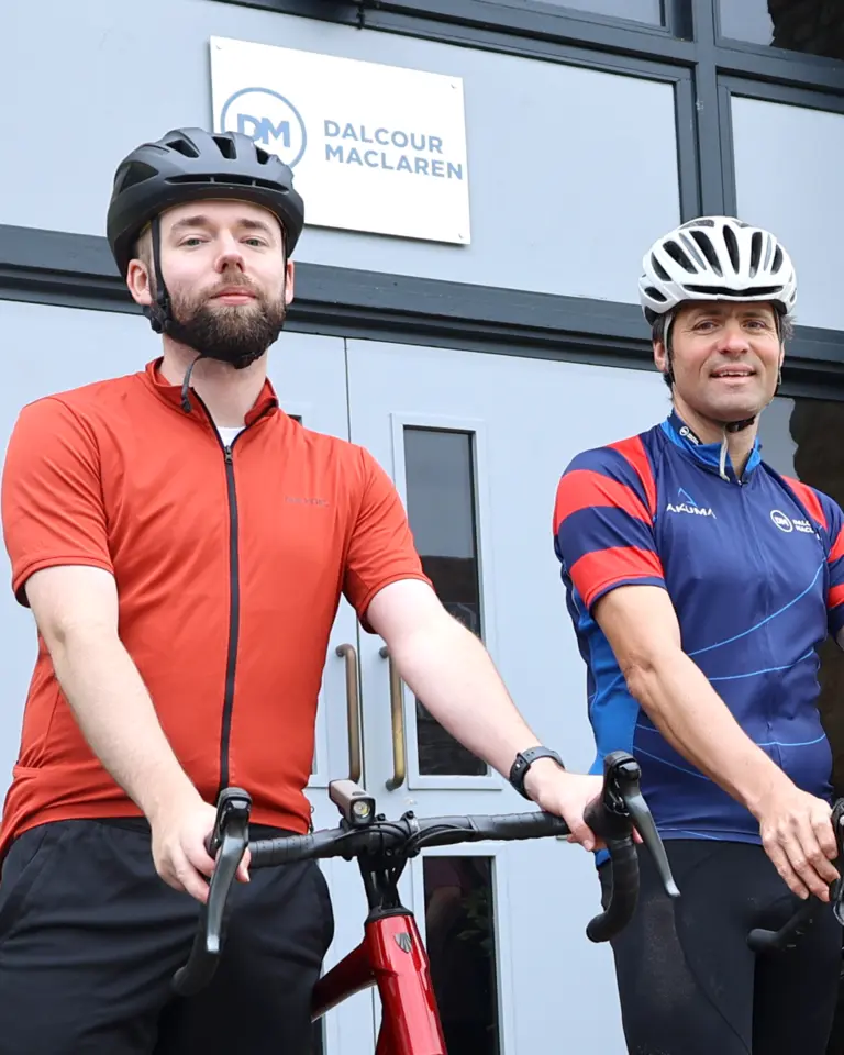 Two men in cycling gear standing with their bikes outside a building. One man wears a red shirt and black shorts, while the other wears a blue and red jersey. Both wear cycling helmets and are posing in front of a glass door with a logo.