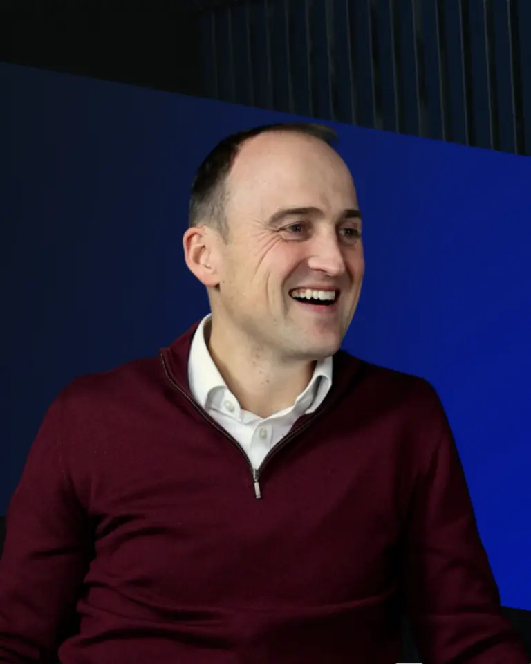 A man in a burgundy sweater smiling while sitting in front of a dark background with blue geometric shapes.