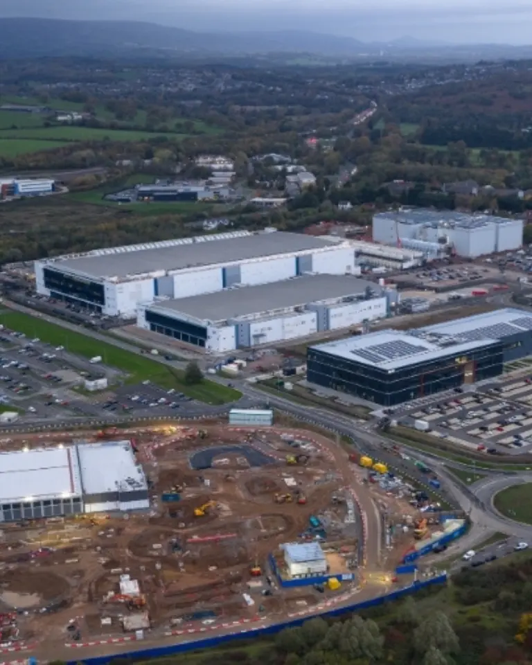 An aerial view of an industrial area featuring several large buildings, construction sites, and parking lots. Surrounding landscape includes green fields and tree lines in the background under a cloudy sky.