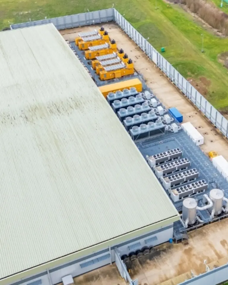 An aerial view of a large industrial building with a green metal roof. Adjacent to the building, various large equipment and generators are arranged on a concrete surface, surrounded by a security fence and grassy areas.