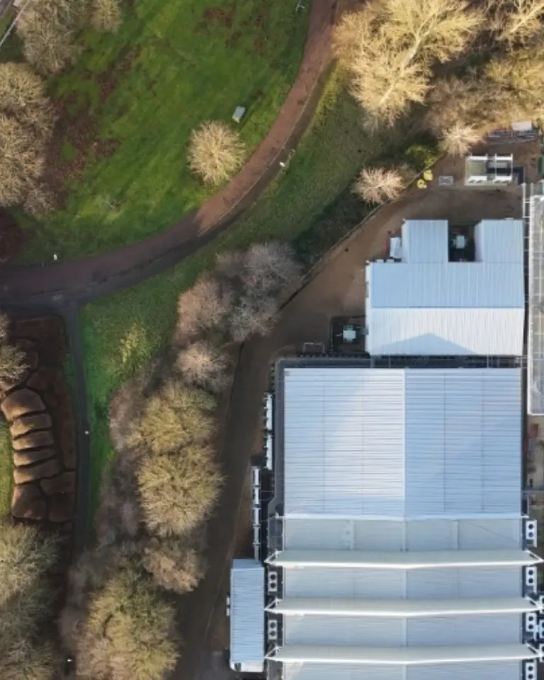 An aerial view of a large building with a flat roof and several ventilation units on top, surrounded by trees. Nearby, there are winding roads and patches of green grass, indicating a rural or suburban setting.