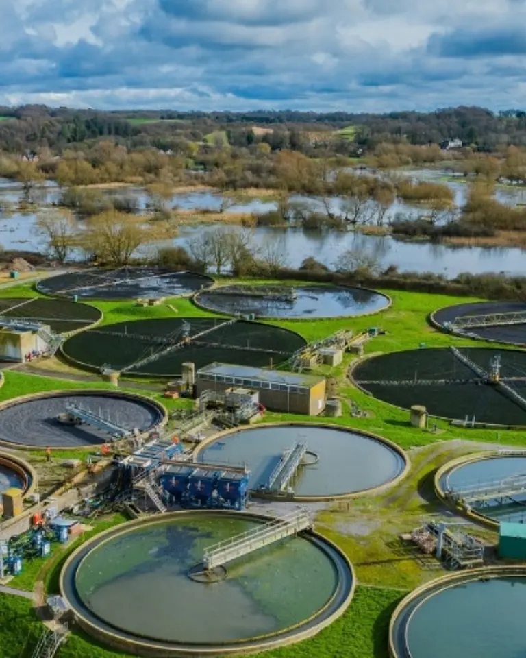 An aerial view of a wastewater treatment facility featuring multiple circular tanks surrounded by green grass. In the background, a river can be seen, along with trees and fields under a cloudy sky.