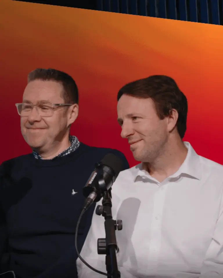Two men stand beside a microphone in front of a red and orange gradient screen, with one wearing a navy sweater and the other a white shirt.
