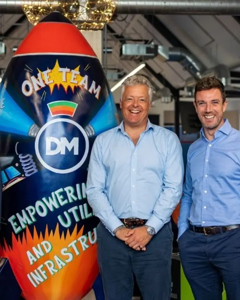 Three men in business attire standing beside a colorful rocket-shaped display with the words "One Team" and "DM" in an industrial office space.