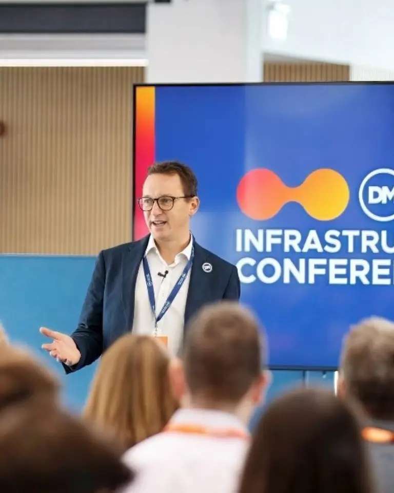 A man in a blazer speaks at the Dalcour Maclaren Infrastructure Conference 26, with an audience seated in the foreground and a presentation screen behind him.