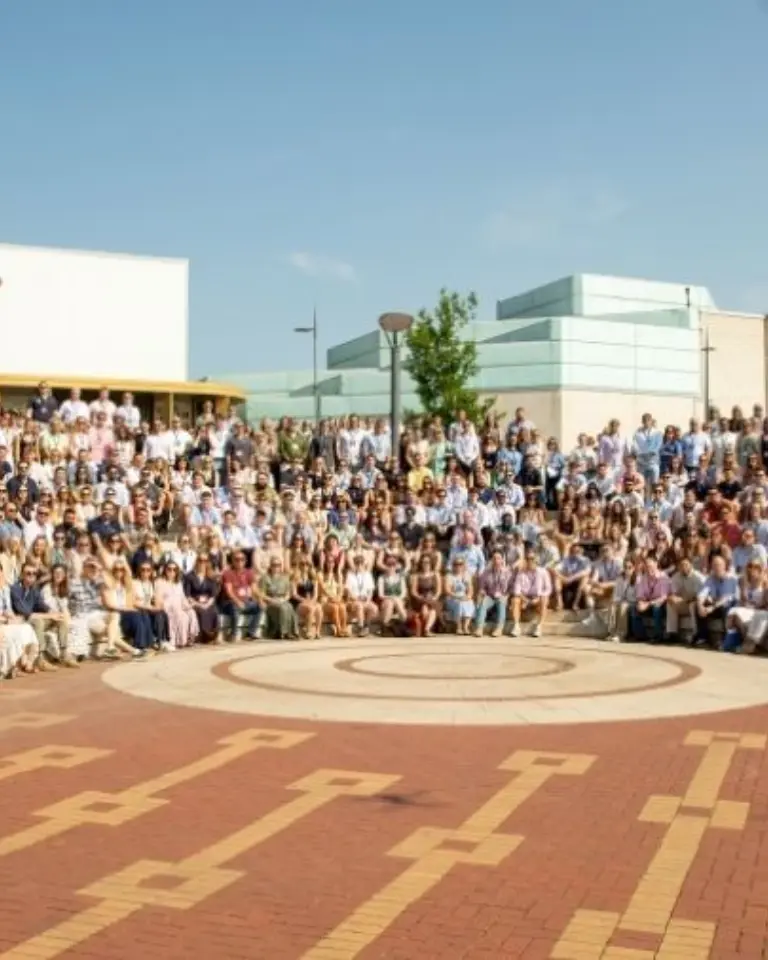 A large group of people poses outdoors on a brick plaza with a circular pattern, in front of modern campus buildings and trees.