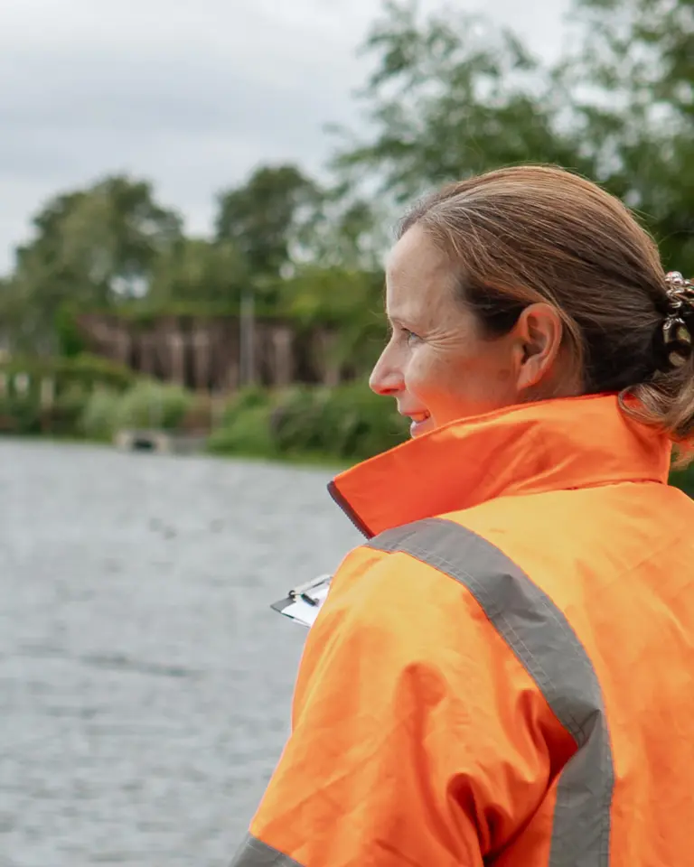Two individuals in bright orange safety jackets standing by a riverbank, observing the water. The setting includes greenery and a cloudy sky.