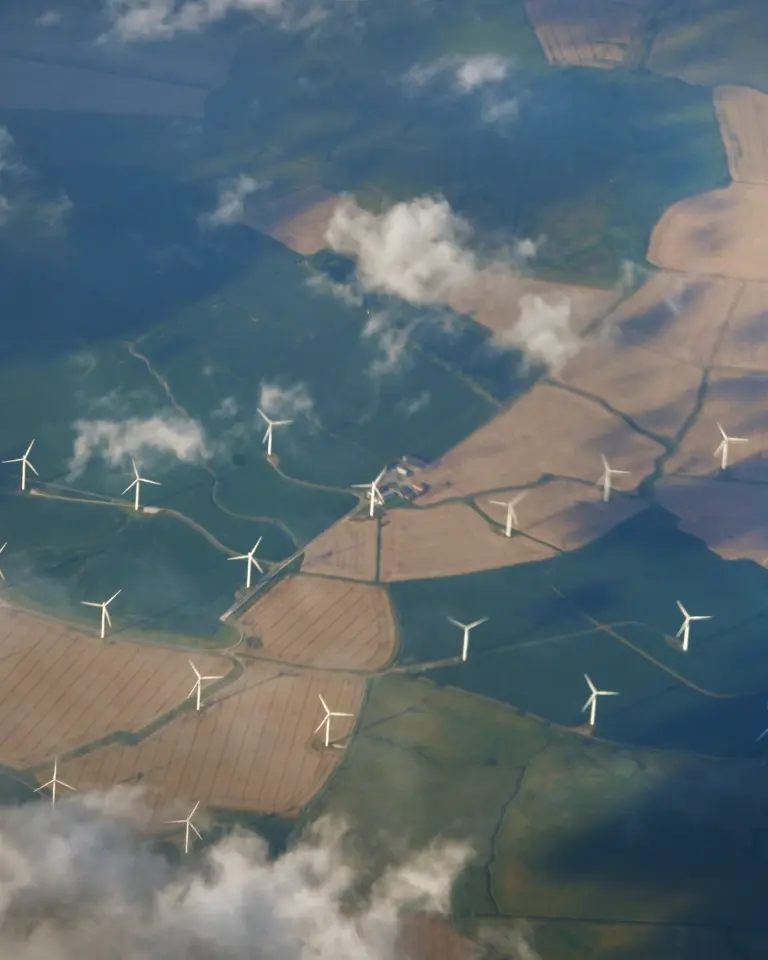 Aerial view of farmland with wind turbines scattered across the fields, under a partly cloudy sky. The landscape features patches of different colors indicating various crops.