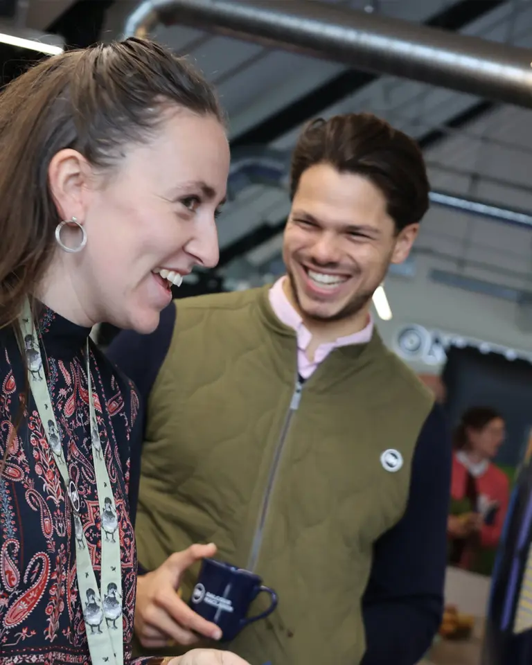 Two smiling individuals engaged in conversation at a lively event. The woman, wearing a patterned blouse, holds a small object while the man, dressed in a green vest, appears amused. A vibrant display with the text 'WE CHALLENGE' is visible in the background.