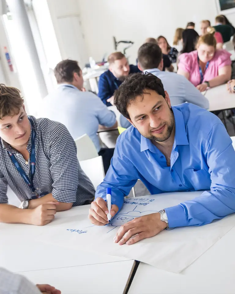 A group of people engaged in a collaborative brainstorming session at a table, with one person writing on a large sheet of paper. Others are observing and discussing ideas, while a presentation screen is visible in the background.