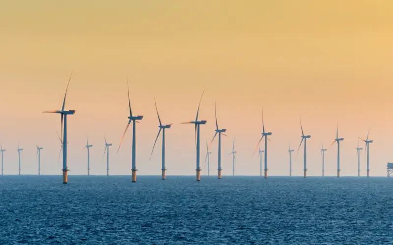 A series of wind turbines lined up in the ocean, with the sun setting in the background, creating a gradient of colors in the sky. The water is calm, reflecting the turbines.