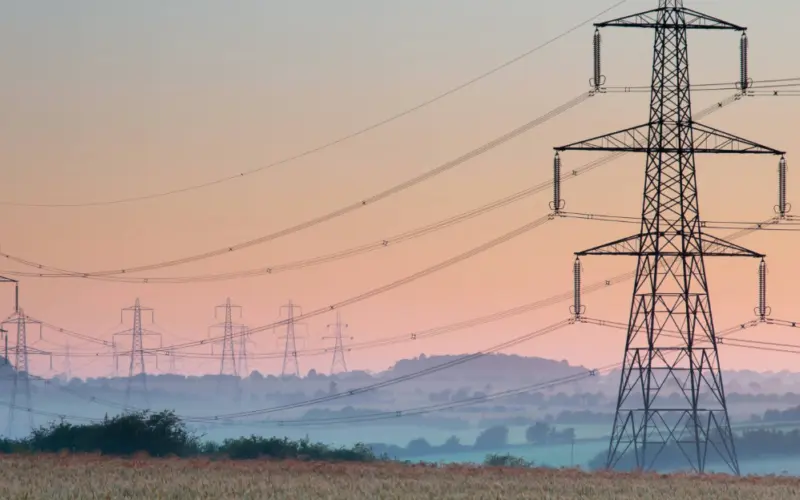 Multiple electricity pylons in a rural landscape at sunrise, with fog covering the fields and distant hills under a soft pastel sky.