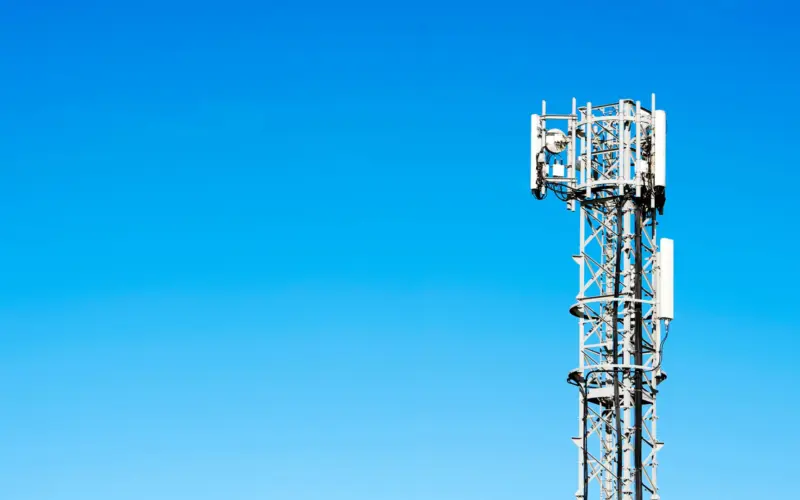 A telecommunications tower against a clear blue sky. The tower features various antennas and equipment mounted on a metal structure.