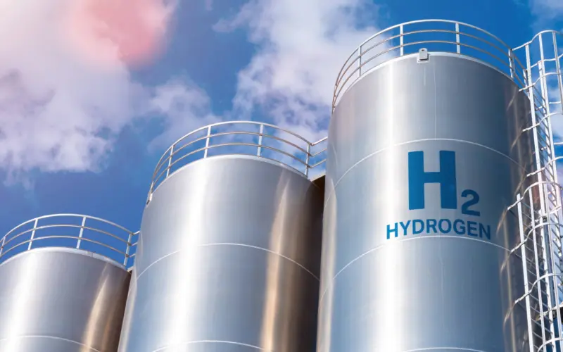Several large metallic tanks labeled 'H2' and 'HYDROGEN' against a clear blue sky, with some clouds visible.