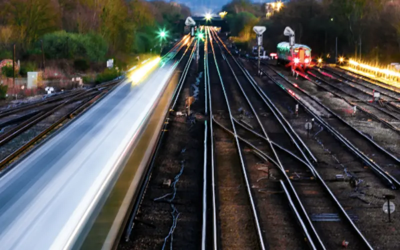 A view of railway tracks at dusk, with train lights creating streaks of light. Multiple tracks curve away into the distance, surrounded by trees and signaling lights.