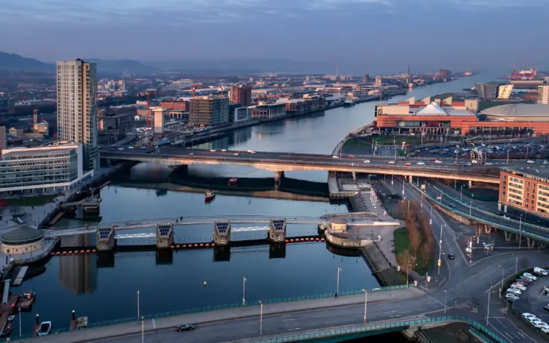 Aerial view of an urban area featuring a wide river, modern buildings, and bridges. A marina and parking lot are visible, along with cityscape in the background. The scene is bathed in early morning light.