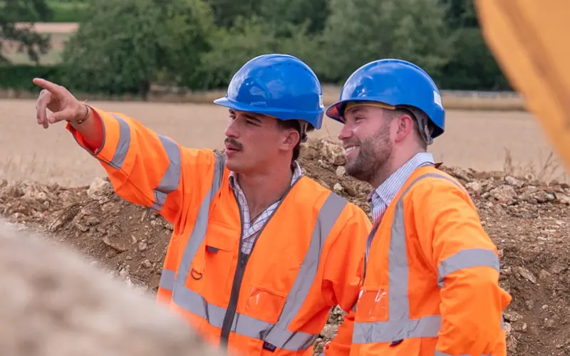 Two construction workers in orange safety vests and blue hard hats discussing a project. One is pointing towards the distance while the other listens attentively, with a rural landscape in the background.