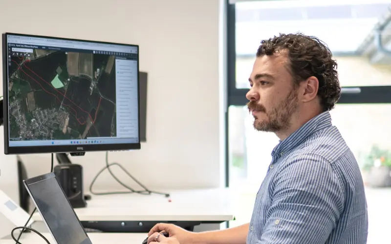 A man sitting at a desk working on a computer, focused on a large monitor displaying a digital map. The workspace is bright with natural light coming from a window.