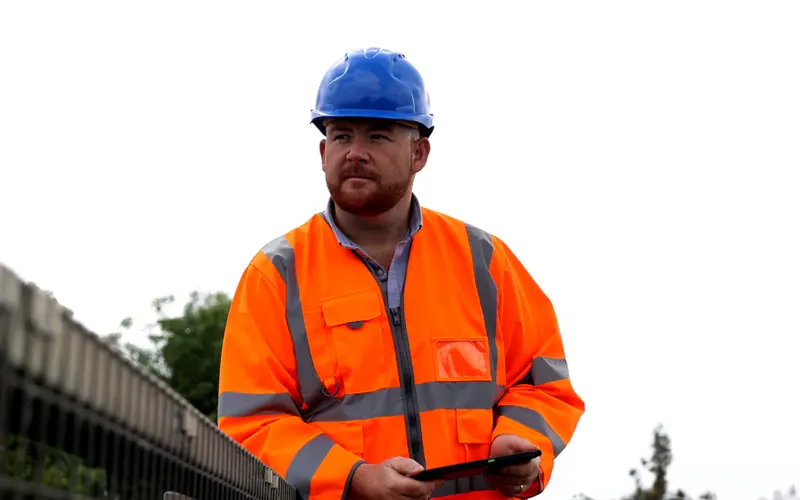 A man wearing an orange safety jacket and a blue hard hat stands near a construction site, holding a tablet. The sky is overcast with trees visible in the background.