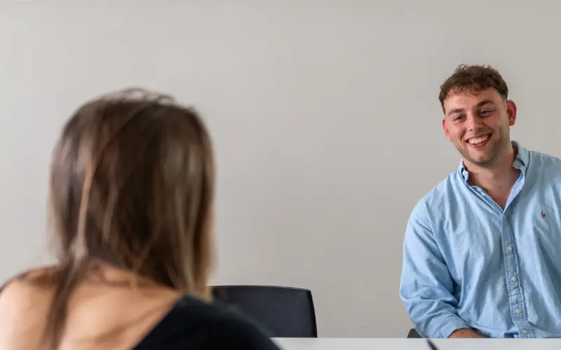 A man wearing a light blue shirt smiling while sitting at a table, facing a woman with long hair, who is turned away from the camera. The background is a simple, minimalistic office space.