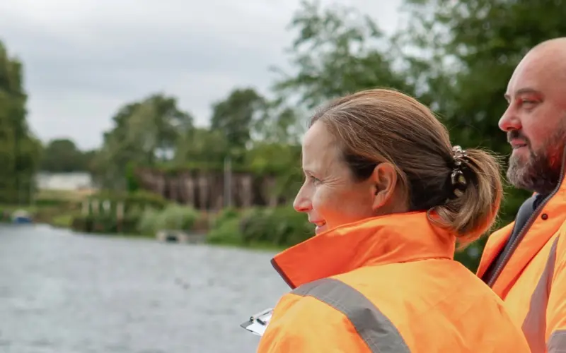 Two individuals wearing bright orange safety jackets are standing by a river, looking at the water. The background features greenery and a cloudy sky, creating a serene outdoor setting.