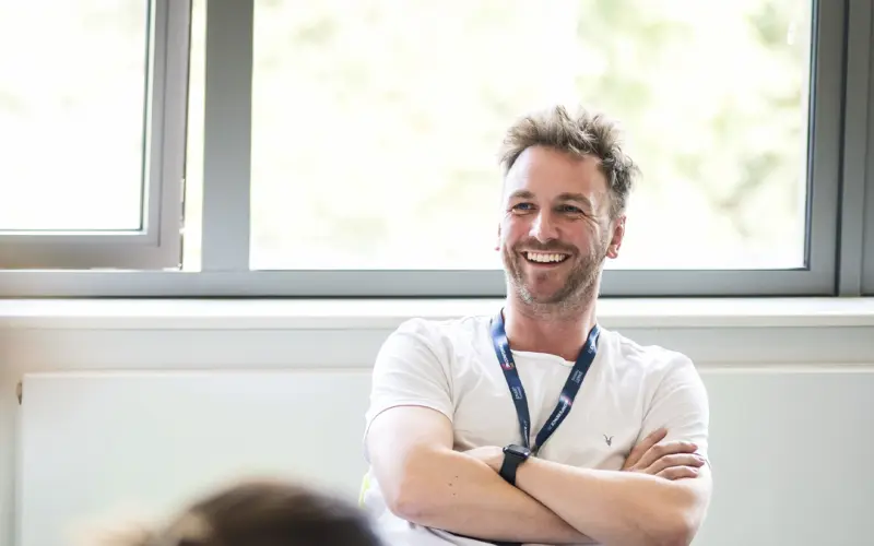 A man with short, light hair wearing a white t-shirt and a lanyard smiles while sitting in a bright room with large windows. The focus is on him, with some blurred figures in the foreground.
