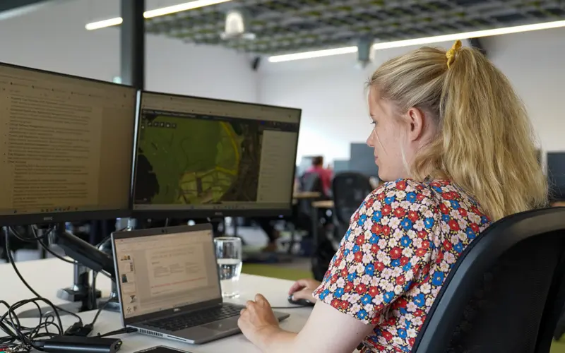 A woman with blonde hair sits at a desk in an office, using a laptop and two external monitors. The screens display text and a map. A glass of water is on the desk.