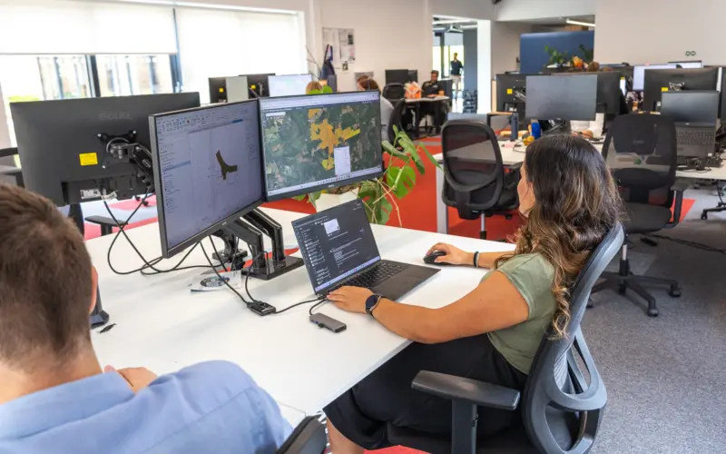 A woman working at a desk with multiple computer monitors in a modern office. She is using a laptop, while a colleague sits nearby. The workspace is filled with other employees and various monitors.