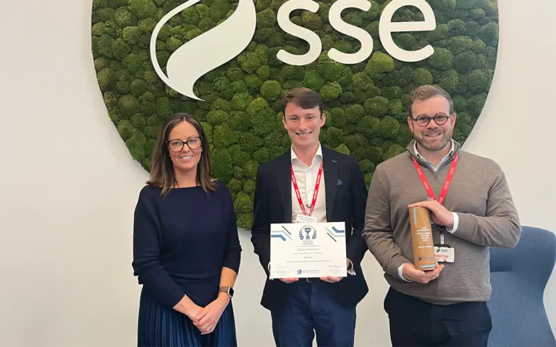A group of three individuals standing in front of a green moss wall with the SSE logo. The woman on the left is wearing a navy sweater, the man in the middle holds a certificate, and the man on the right is holding a wooden award.