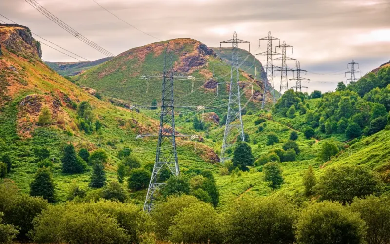 A scenic landscape featuring rolling hills covered with greenery, electric power lines towering in the background, and a cloudy sky. The foreground includes clusters of trees and bushes.