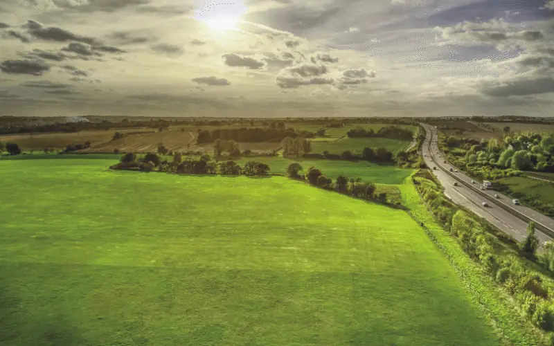Aerial view of a lush green field bordered by trees, with a highway running alongside it. The sky is partly cloudy, and sunlight is visible, casting a warm glow over the landscape.