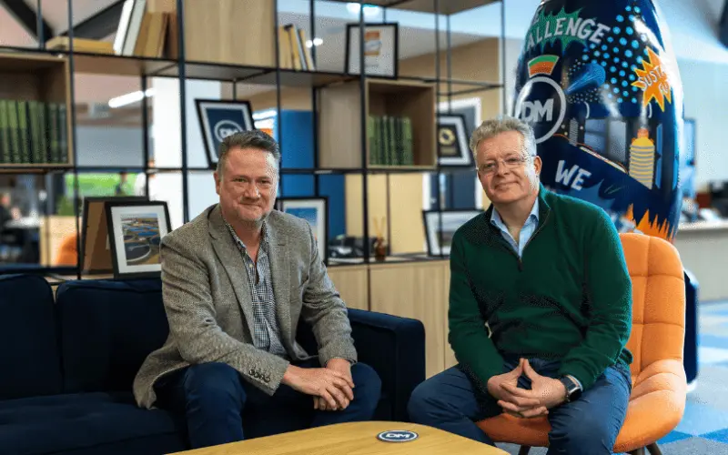 Two men seated on a couch in a modern office setting. One man is wearing a blazer and the other a green sweater. They are positioned around a wooden coffee table, with books and framed pictures in the background.