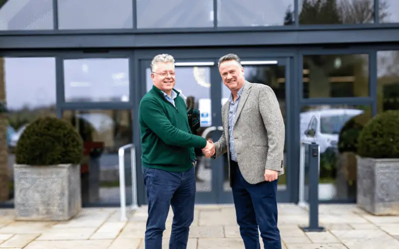 Two men shaking hands in front of a modern building entrance. One man is wearing a green sweater and the other is in a gray blazer. Large glass doors and planters are visible in the background.