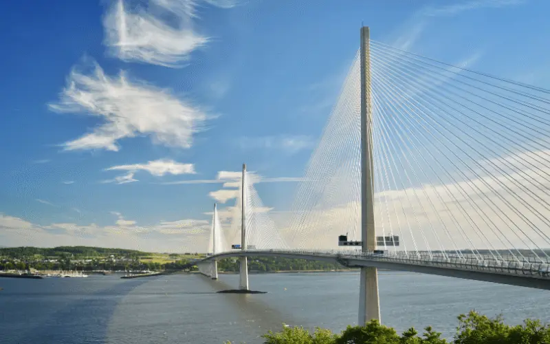 A suspension bridge with tall towers and cable stays spans across a river, beneath a clear blue sky with scattered clouds. Lush greenery is visible in the foreground.