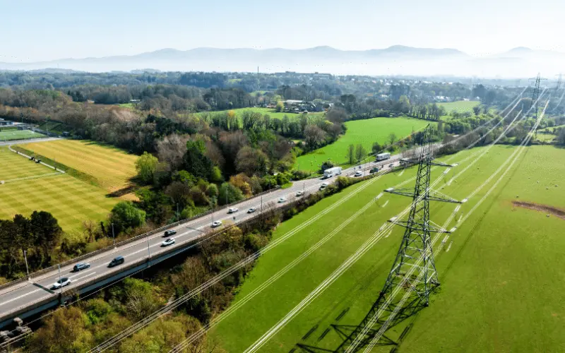 An aerial view of a rural landscape with green fields, a highway with cars, and power lines. Rolling hills are visible in the background under a clear blue sky.