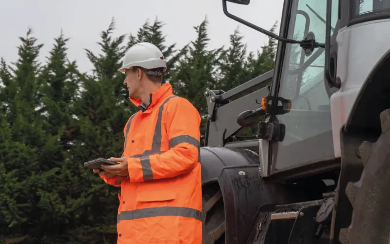 A construction worker in an orange safety jacket and white hard hat stands beside a large machinery, holding a tablet while looking at it. Green trees are in the background.