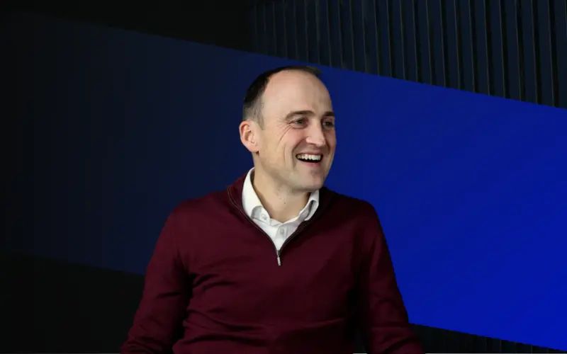 A man in a burgundy sweater smiling while sitting in front of a dark background with blue geometric shapes.