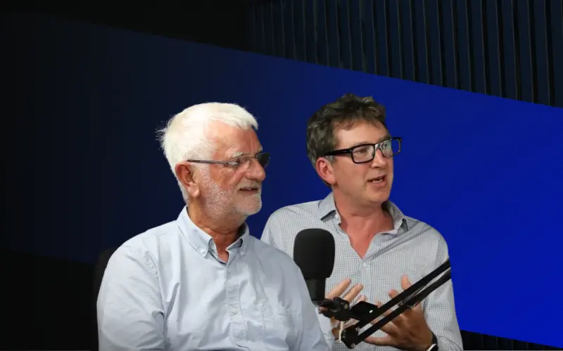 Two men seated at a table with microphones in front of them, engaged in conversation. One man has white hair and glasses, while the other has dark hair and is also wearing glasses. The background features a blue geometric design.