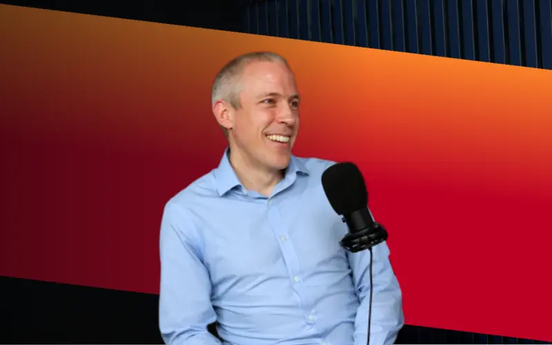 A man in a light blue shirt smiling while sitting next to a microphone, against a gradient background transitioning from orange to dark blue.