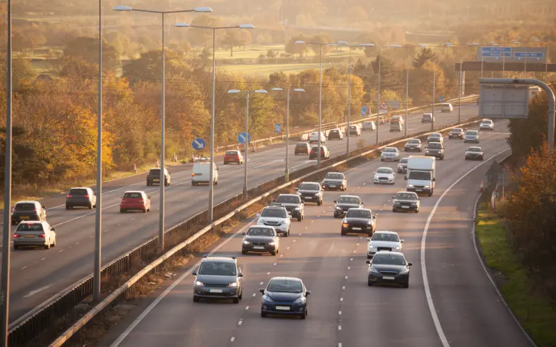A view of a multi-lane highway with several vehicles, including cars and a truck, traveling in both directions. The scene is set during sunset, with trees and fields visible in the background.