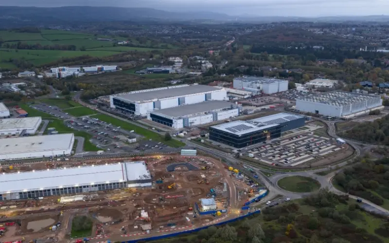 An aerial view of an industrial area featuring several large buildings, construction sites, and parking lots. Surrounding landscape includes green fields and tree lines in the background under a cloudy sky.