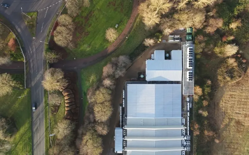 An aerial view of a large building with a flat roof and several ventilation units on top, surrounded by trees. Nearby, there are winding roads and patches of green grass, indicating a rural or suburban setting.