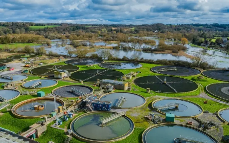 An aerial view of a wastewater treatment facility featuring multiple circular tanks surrounded by green grass. In the background, a river can be seen, along with trees and fields under a cloudy sky.
