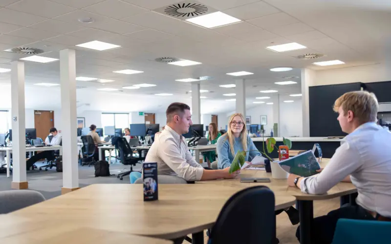 Three office workers sit around a wooden table in an open-plan office, with desks, computers, and large wall monitors in the background.